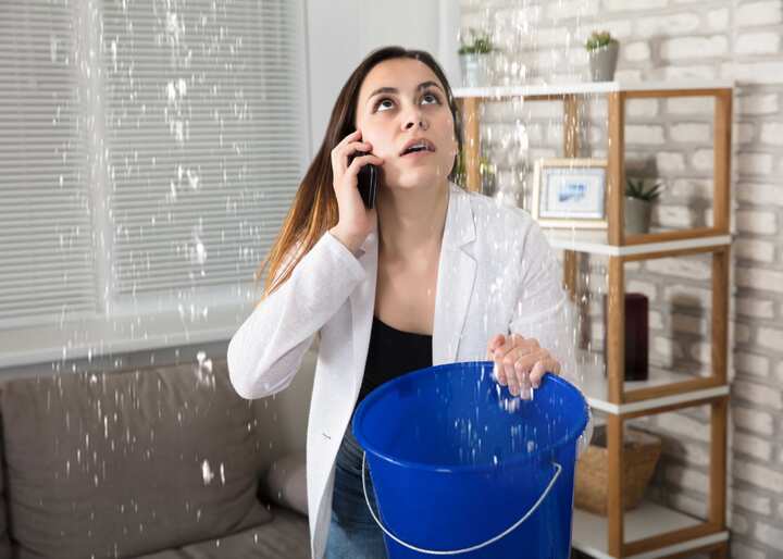 A woman calling water damage repair experts while holding a bucket trying to catch the water leaking from her ceiling in Manchester Township